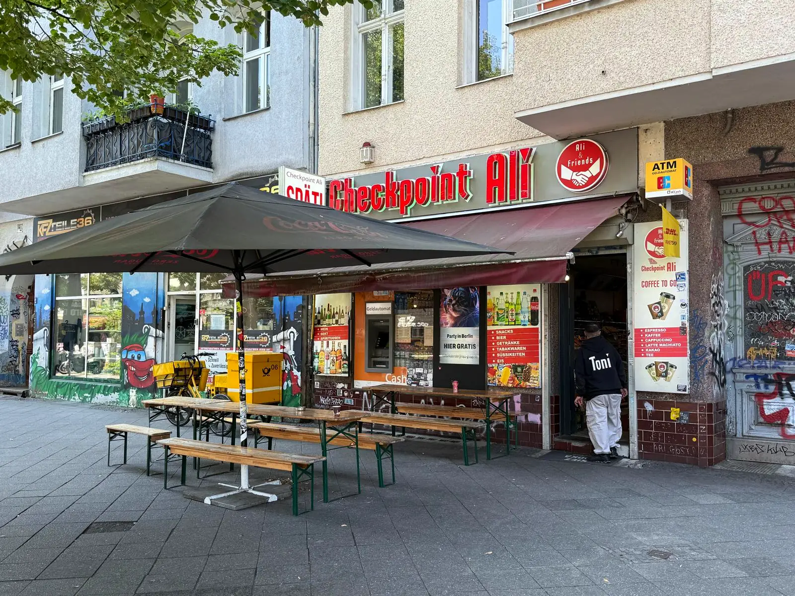 Außenansicht von Checkpoint Ali in der Wiener Straße in Berlin-Kreuzberg mit Biertischen und Sonnenschirm vor dem Laden